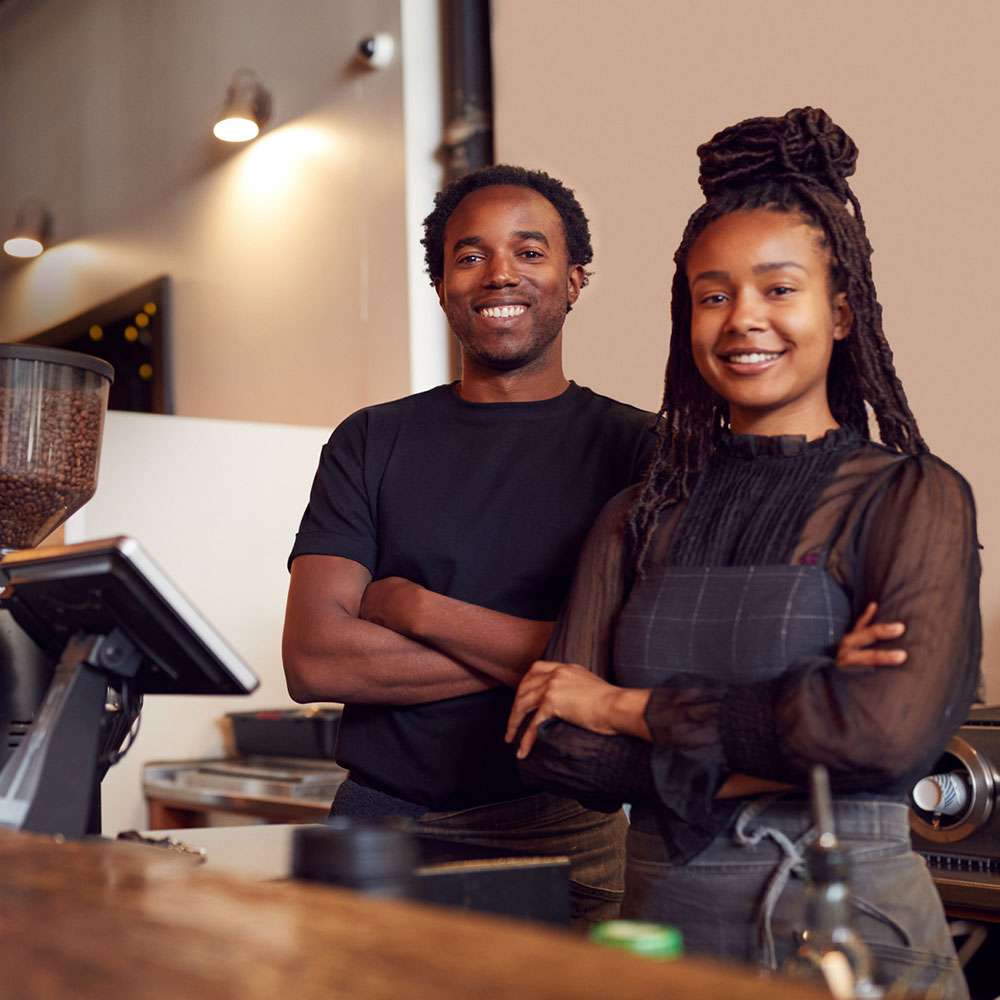 Two African American Business Owners stand behind the counter at their coffee shop.